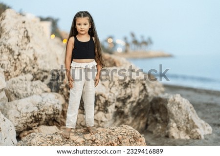 Full length view of the little girl wearing black top and beige pants standing on rocks while posing on the beach. Travel vacation, summer concept