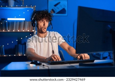 Young arabic gamer wearing headphones playing video game on personal computer, play in eSport cyber games tournament in neon light. Funny teenager is excited because of seen virtual video. Copy space