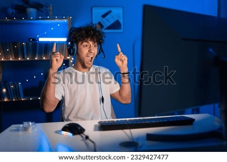 Surprised young arabic gamer wearing headphones playing video game on personal computer, looks at camera with a shocked face, play in eSport cyber games tournament in neon light. Copy space Royalty-Free Stock Photo #2329427477