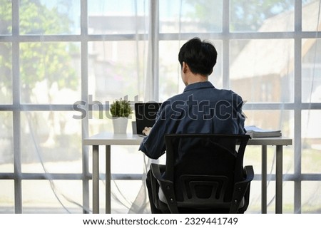 Back view image of a focused Asian businessman working on his work on his laptop computer at his desk by the window in a modern office. Royalty-Free Stock Photo #2329441749