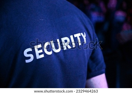 The security guard wears a T-shirt with the inscription security. A security guard stands in front of the audience during a music festival. Royalty-Free Stock Photo #2329464263