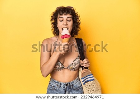 Curly-haired woman with beach gear enjoying ice-cream, yellow studio backdrop.