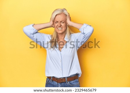 Middle-aged Caucasian woman in blue shirt, yellow studio covering ears with hands trying not to hear too loud sound. Royalty-Free Stock Photo #2329465679