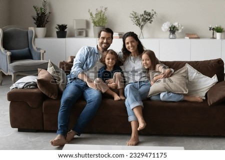Portrait of smiling young multiracial family with two small kids sit relax on couch in living room. Happy multiethnic parents renters with little children rest at home together. Rental concept.