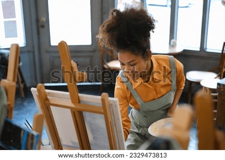 African American female artist wearing apron painting picture with brush on easel in art studio