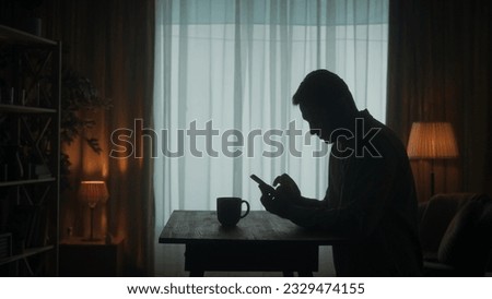 A man stands at a table with a phone in his hands, a cup of coffee stands nearby. A man types a message, looks at photos, videos. The dark silhouette of a man in the living room. Royalty-Free Stock Photo #2329474155