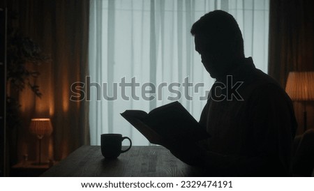 A man reads a book sitting at a table with a cup of coffee, tea in the room on the background of the window. Dark silhouette of a man with an open book in his hand close up. Royalty-Free Stock Photo #2329474191