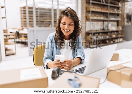 Online store owner reading a text message on her cellphone. Happy businesswoman making plans for product shipping in her warehouse. Female entrepreneur running an e-commerce small business. Royalty-Free Stock Photo #2329485521