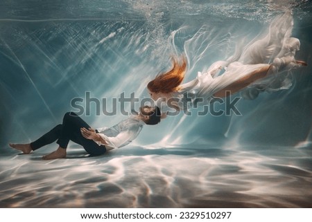 Happy couple in love swim underwater, female goddess muse inspires male writer poet creator. Nymph girl saves drowning guy at bottom sea under water. Red hair white long silk dress float. Art photo.
