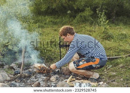 A young red-haired man lights a fire in a tourist parking lot on a summer day in sunny weather. High quality photo