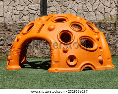 Orange cave toy, with holes. Kids playground. In the park. Stone wall, background