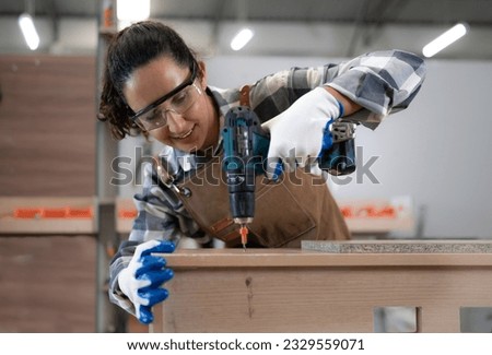 Beautiful Latin woman carpenter using power tools working her wood job in carpenter's shop. Young hispanic female in protective goggles busy in furniture woodworking. Feminism in carpentry industry. Royalty-Free Stock Photo #2329559071