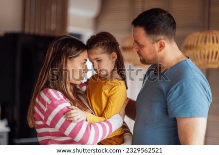 Young parents comfort their cute daughter. Beautiful woman hugs and talks to little girl in living room at home. Dad and mom take care of their child. Tender moment family support and understanding. Royalty-Free Stock Photo #2329562521