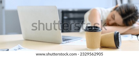 Two coffee paper cups on table with a tired business Asian woman sleeping in the background. working overtime concept. Select focus to paper cup. Banner cover design