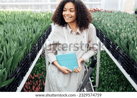 Young cute african girl is using digital tablet to work with tulips in greenhouse. Darkskinned female farmer stands on stepladder among grown flowers. Modern technology in gardening business.