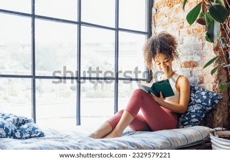 Pretty african american girl in homewear, reading book sitting on windowsill. Beautiful Afro woman happily reading a novel in cozy room. Education and knowledge concept Royalty-Free Stock Photo #2329579221