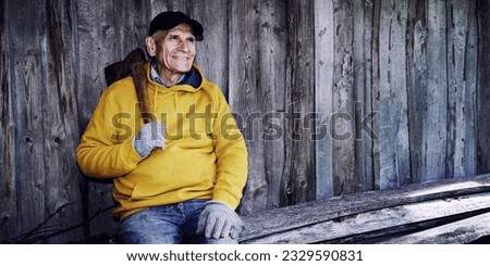 Senior lumberjack with old iron axe resting against wooden rustic barn wall. Mature woodman sitting outdoor. Royalty-Free Stock Photo #2329590831