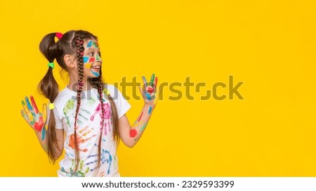 A child smeared with paint. A little girl painted with multicolored paints points to your advertisement. Children's creativity. Yellow isolated background. Copy space. Banner.