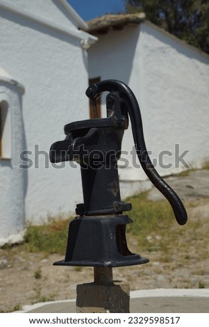 With a bright white building in the background this image of a black water pump with a handle in the grounds of an orthodox church in Greece.