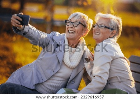 Two older women taking selfies and smiling. They are sitting on a bench in the autumn