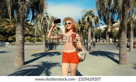Portrait of young woman taking selfie with phone in summer park wearing backpack, straw hat on palm tree background