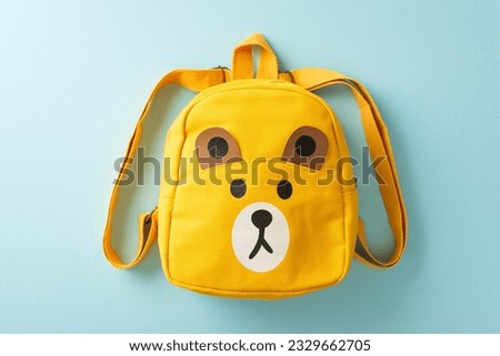 Elementary school education concept. High angle view picture of cute yellow school bag with cartoon bear print for small children on pastel blue isolated background