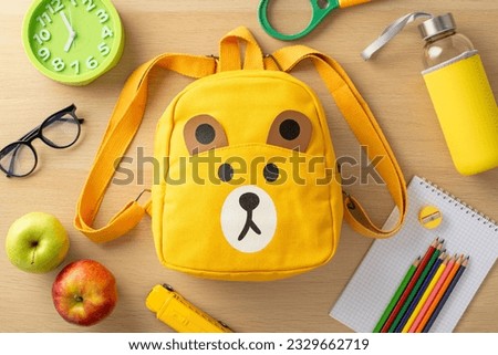 Primary education concept. Top-view image of assorted supplies, notepad, color pencils, sharpener, glasses, clock, toy, magnifier, playful bear-shaped backpack, water bottle, and apples on wooden desk