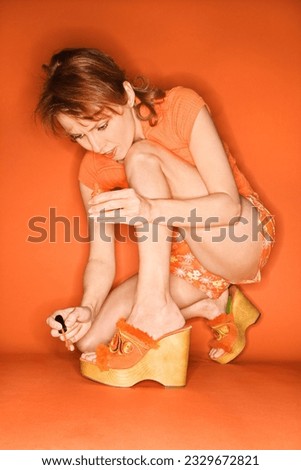 Caucasian mid-adult woman on orange background painting her toenails with nail polish.