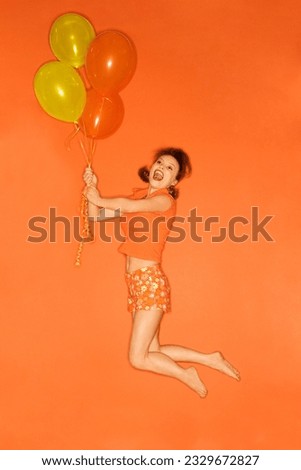 Caucasian mid-adult woman being lifted into the air by balloons, on orange background.