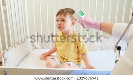 The doctor is using an electronic thermometer to measure the child's temperature who is sitting on the bed. Kid aged about two years (one year eleven months)