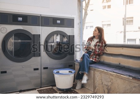 Young Woman In Casual Doing Laundry And Using Cell Phone, Sitting Waiting In Laundrette Room Near Washing Machines. Lady Booking Laundromat Via Mobile Application Posing Indoors Royalty-Free Stock Photo #2329749293