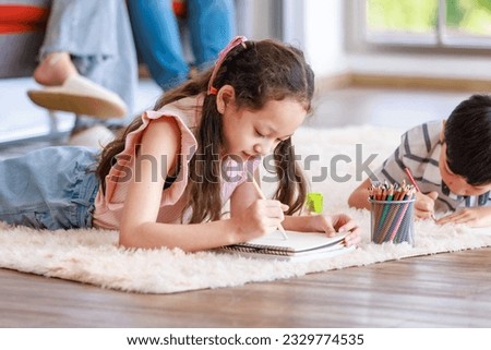 Closeup Asian happy cheerful joyful family little boy son and girl daughter laydown on carpet floor helping teaching painting drawing cartoon with color pencils in living room.