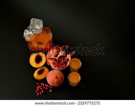 Two glasses and a glass decanter with homemade liqueur on a black background, next to pieces of a ripe peach and a broken pomegranate. Top view, flat lay.