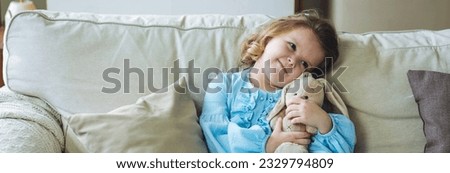 Cute little girl in blue dress on the sofa at home with her favourite toy, teddy rabbit. Concept of children playing. Banner Royalty-Free Stock Photo #2329794809