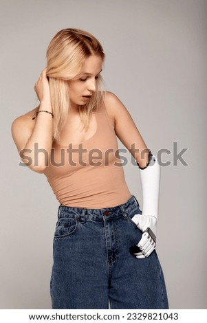 Studio half-length portrait of beautiful young blonde girl with disability wearing sensory bionic prosthetic arm. Modern woman freely controls artificial robotic hand. Full life after limb loss. Royalty-Free Stock Photo #2329821043