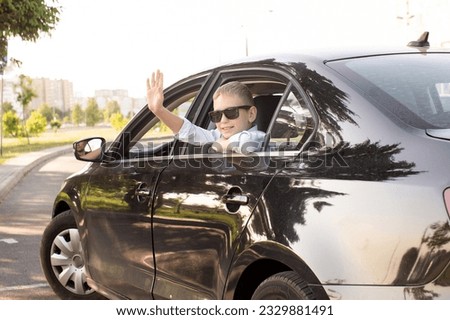 A cute boy climbed out of the car window wearing sunglasses and laughing. Waving to friends. Contour light. Saying goodbye before leaving, on the move, going on vacation Royalty-Free Stock Photo #2329881491