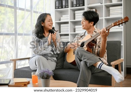 A happy and smiling Asian grandmother is clasping her hands while listening to her grandson sing and play guitar, spending quality time at home together. Royalty-Free Stock Photo #2329904019
