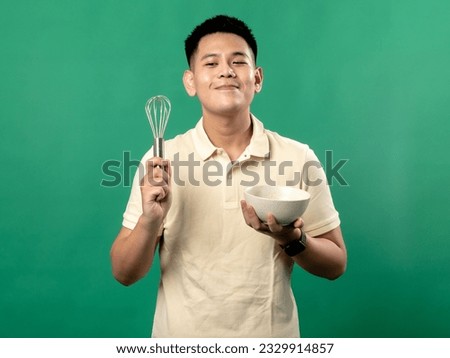 A portrait of an Indonesian Asian man wearing a yellow shirt, looking happy while posing and whisking eggs in a bowl, isolated on a green background.