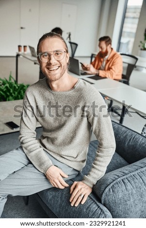 happy entrepreneur in stylish eyeglasses looking at camera in modern coworking environment near colleagues working on blurred background, successful entrepreneurship concept