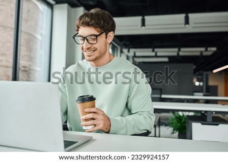 smiling businessman in casual clothes and stylish eyeglasses holding paper cup with takeaway drink and working on laptop in modern office environment, successful business concept Royalty-Free Stock Photo #2329924157
