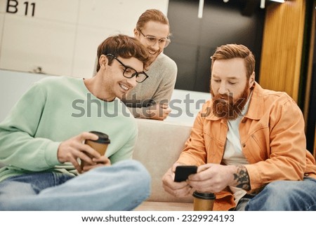 happy businessmen in eyeglasses looking at bearded colleague browsing internet on mobile phone on comfortable couch, coffee break in lounge of modern coworking space