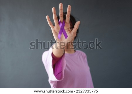 Woman in purple t-shirt holding showing purple awareness ribbon on palm, covering face. Symbol is used to raise awareness for Alzheimer's disease, domestic abuse, cancer disease Royalty-Free Stock Photo #2329933775