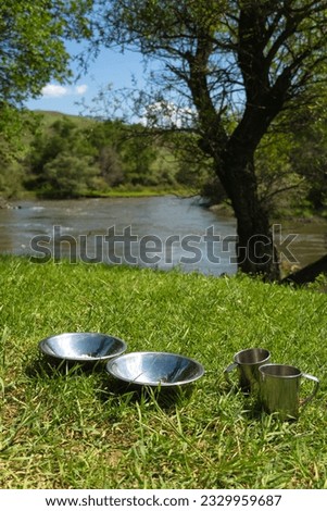 Close-up of aluminum plates and mugs stand on the green grass against the incredible landscape of a mountain river and trees on a salty day. Cooking while traveling by car. Vertical photo