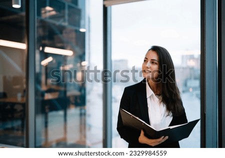Young smiling female entrepreneur in formal wear standing with documents near panoramic window during work in contemporary company looking away Royalty-Free Stock Photo #2329984559