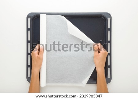 Young adult woman hand holding white roll of baking paper and covering dark black oven tray on light gray table background. Closeup. Point of view shot. Royalty-Free Stock Photo #2329985347