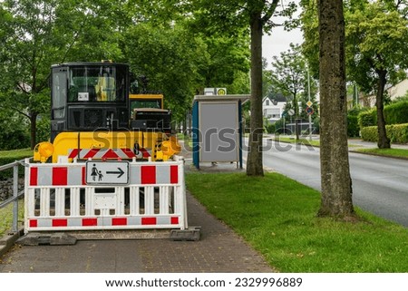 A yellow excavator fenced with plastic in white and red barrier shields stands on the sidewalk.