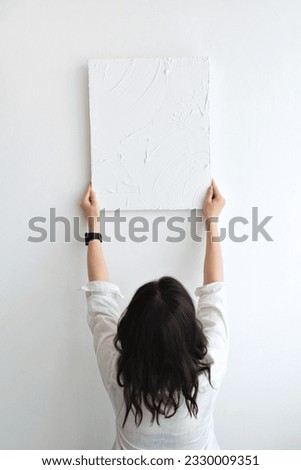 Young brunette woman holding picture canvas with abstract plaster artwork on a white wall background. Minimalist aesthetic artist work design concept