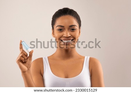 Cheerful beautiful young african american woman wearing white top holding lipstick and smiling, black lady applying hygienic lip balm on light background, copy space