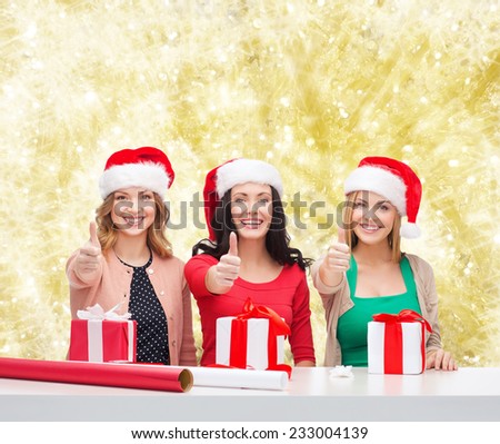 christmas, holidays, celebration, decoration and people concept - smiling women in santa helper hats with decorating paper and gift boxes showing thumbs up over yellow lights background