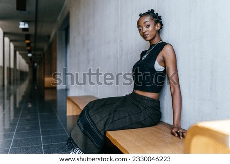 Portriat of african millennial woman posing indoor sitting on bench over gray wall background. Female student sitting in hallway interior at campus while resting after long hard lessons.